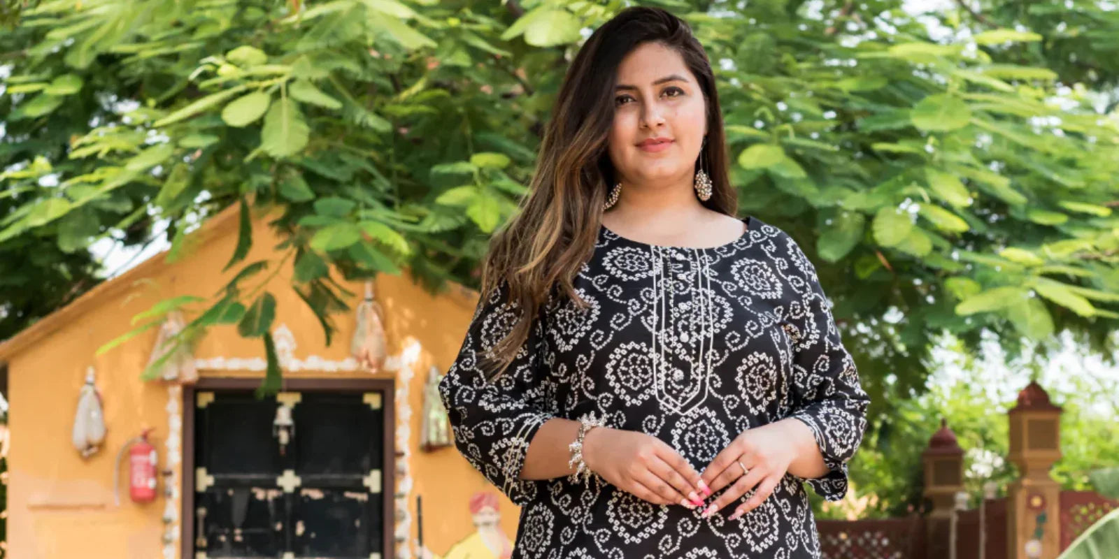 Plus size woman wearing black and white printed kurta posing outdoors near a yellow building with green foliage