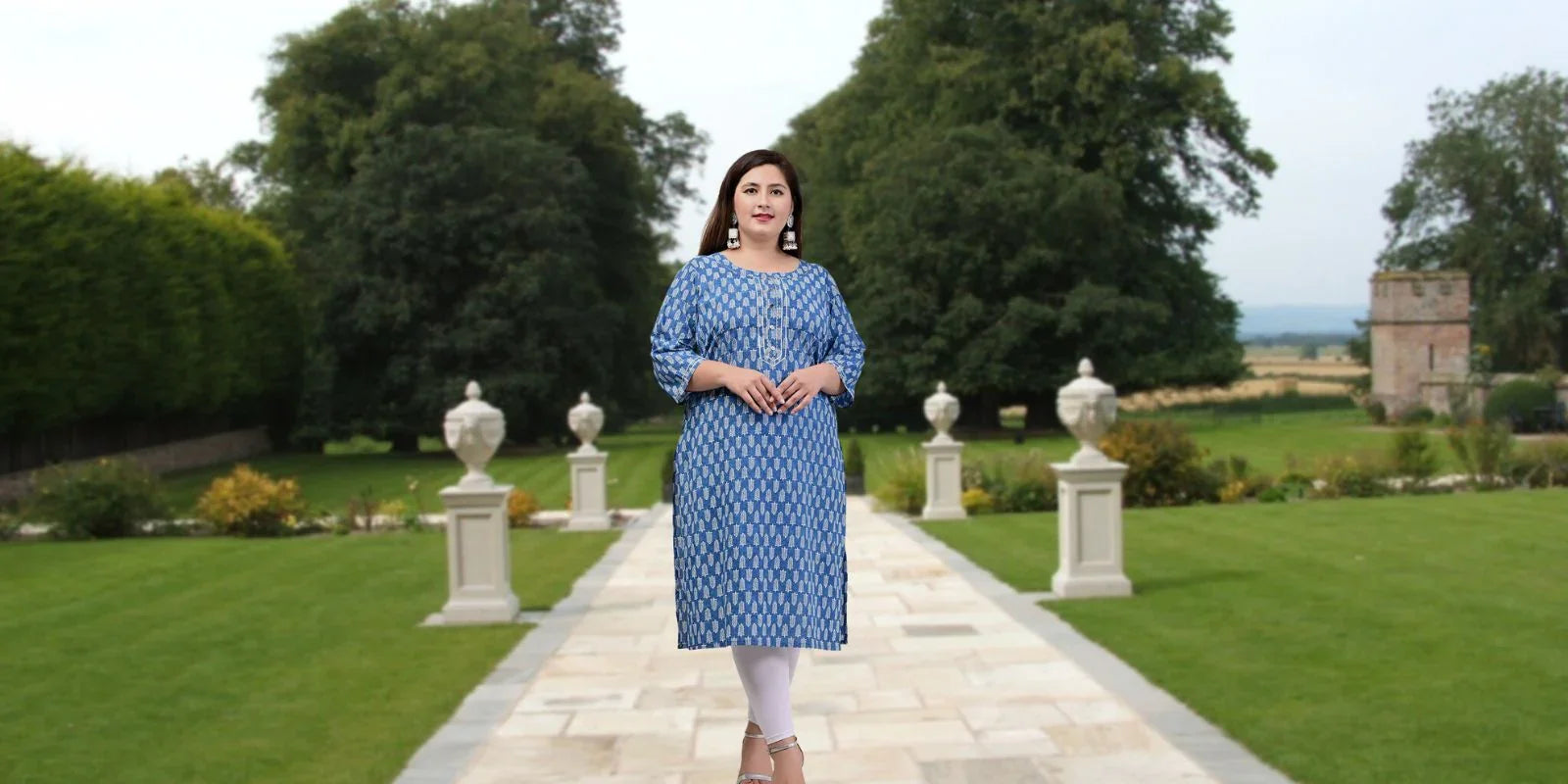 Woman in blue patterned kurti and white leggings standing on garden pathway with green lawns and trees