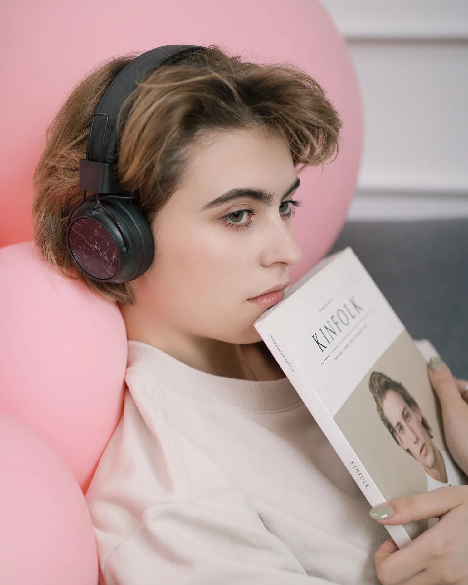 Young person with short hair wearing black headphones, resting on pink balloons, holding a Kinfolk magazine
