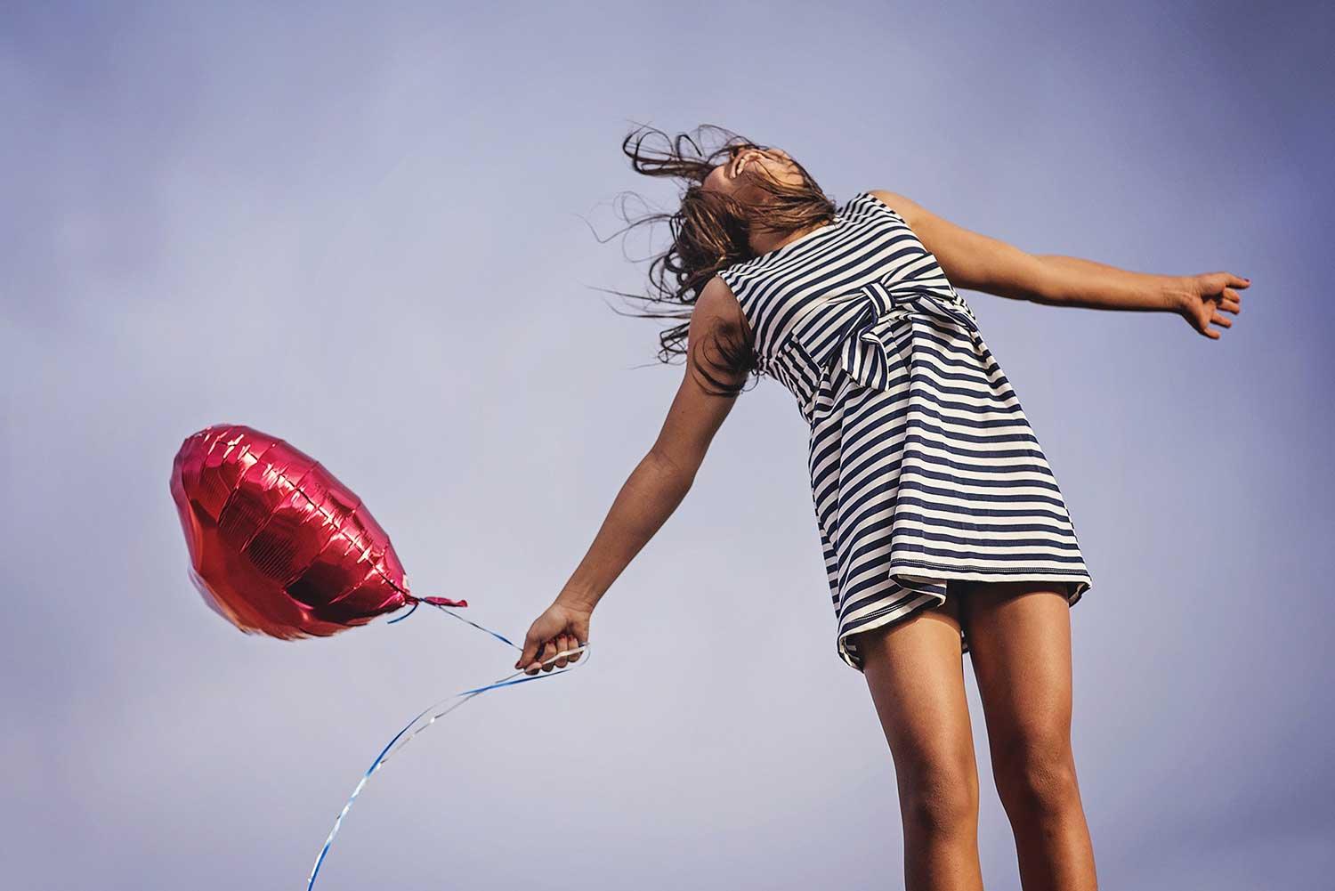 Girl in striped dress holding red heart-shaped balloon against clear sky