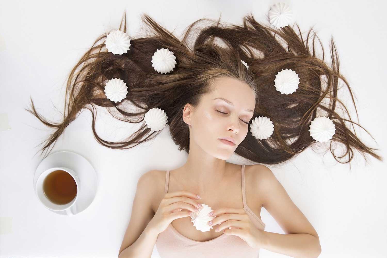 Sleeping woman with long brown hair scattered with white meringue cookies and cup of tea on white background