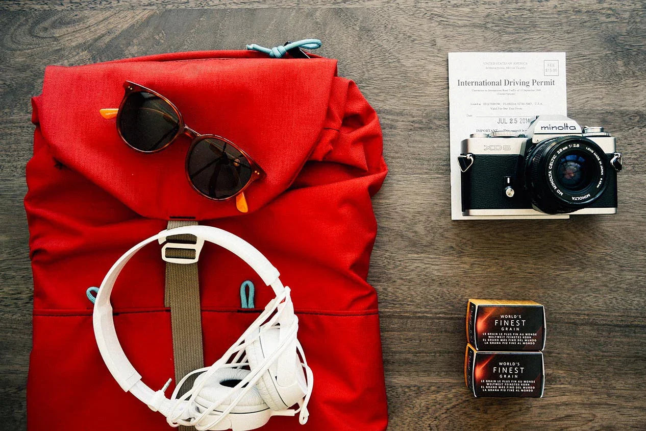 Red backpack with sunglasses and white headphones next to Minolta camera, driving permit, and soap bars on wooden surface