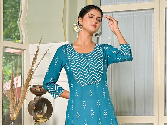 Woman wearing teal kurti with white print standing indoors near a window and decorative lamp