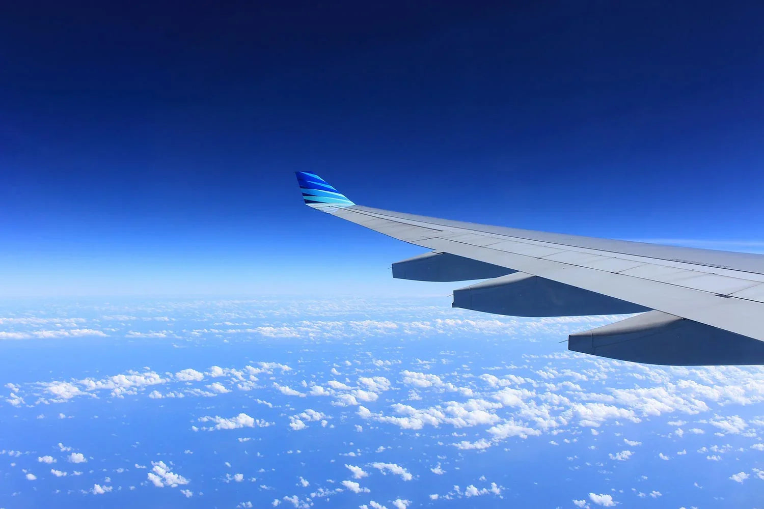 View of airplane wing with blue striped winglet flying above scattered clouds under deep blue sky