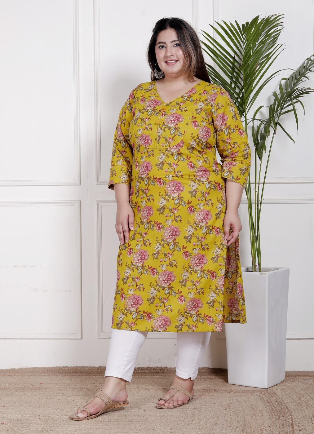 Woman wearing a yellow floral kurta with white pants standing indoors next to a plant.