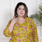 Woman wearing a yellow floral dress against a white wall with a plant.